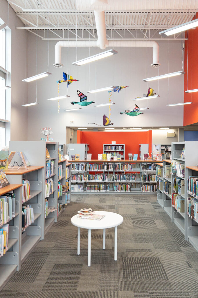 Interior of a contemporary public library's children's section with low bookshelves and kid furniture. The wall to the right is bright red. There are fabric birds hanging from the ceiling.