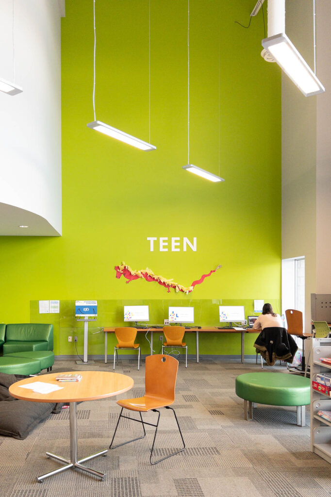 Interior of a contemporary library with a bright green wall on which the word "Teen" is printed. A person is sitting at a row of computers against the wall.