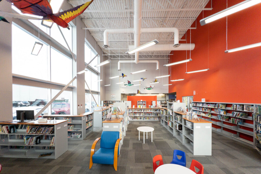 Interior of a contemporary public library's children's section with low bookshelves and kid furniture. The wall to the right is bright red. There are fabric birds hanging from the ceiling.
