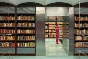 Interior of a contemporary library with stainless steel and glass structures interspersed with wooden bookshelves. Through a doorway, a sculpture representing a pink standing figure can be seen.