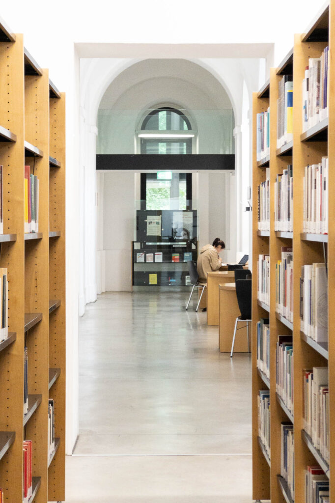 Interior of a refurbished neobaroque building. It is a corridor with vaulted ceilings, seen through rows of wooden bookcases. A person sits at a table working.