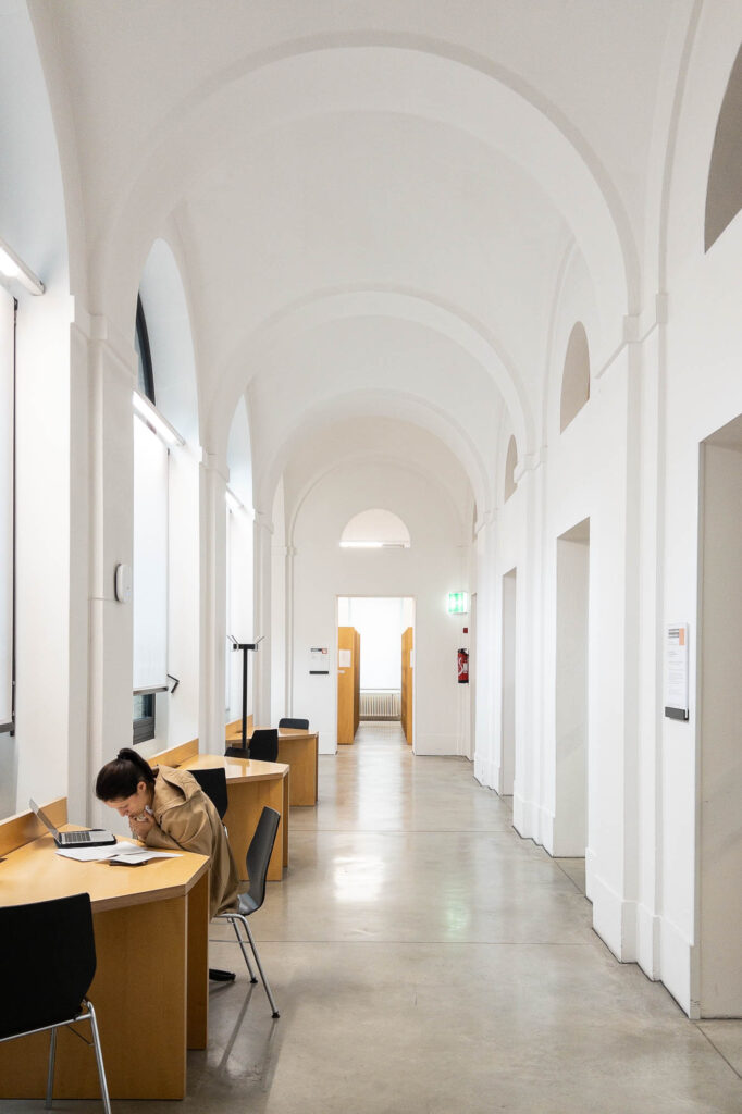 Interior of a refurbished neobaroque building. It is a corridor with vaulted ceilings. To the left, small work tables face the windows, a person occupying the frontmost table. To the right are a series of doors.