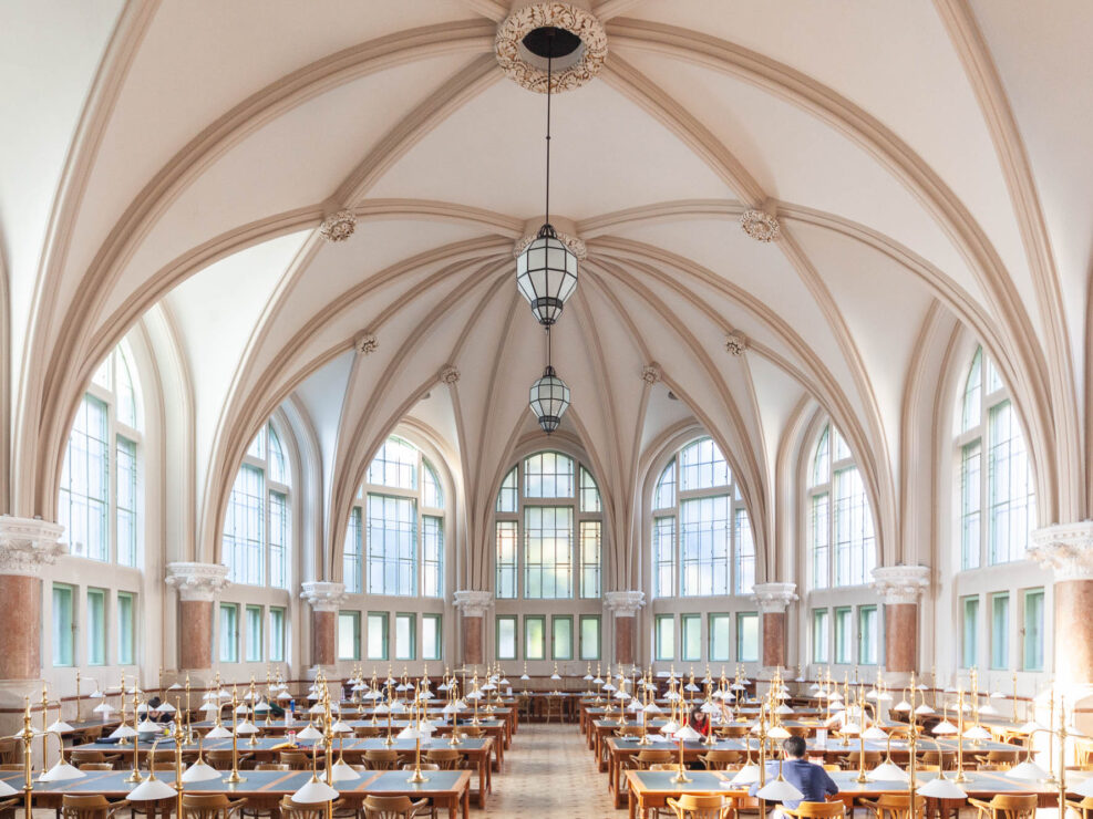 Interior of a Neogothic library reading room, showing a high rib vaulted ceiling and fan windows. Chandeliers hang from the ceiling. The room is filled with long work tables, on which a few students are seated.