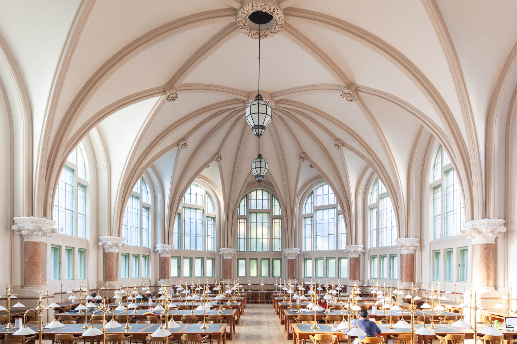 Interior of a Neogothic library reading room, showing a high rib vaulted ceiling and fan windows. Large chandeliers hang from the ceiling. The room is filled with long work tables, on which a few students are seated.
