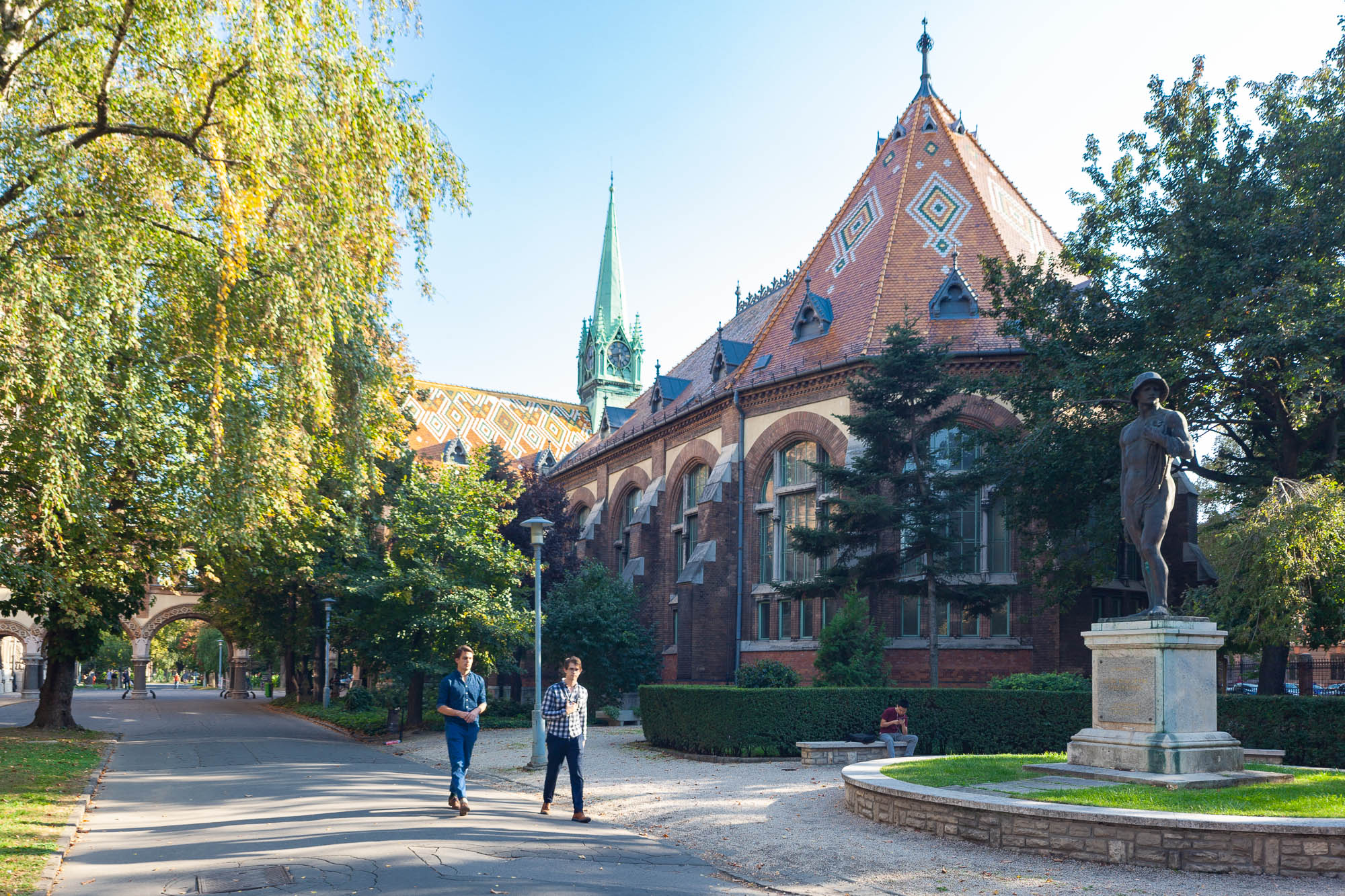 Outside view of the BME-OMIKK library in Budapest. It is a brick building with a colourful tile roof surrounded by a park.