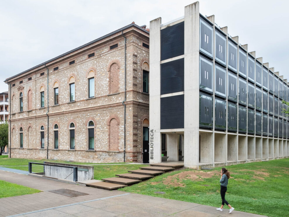 Exterior of the USI library in Lugano, Switzerland. It is a neobaroque building with vaulted windows and unadorned stone facade, with a contemporary concrete and metal extension to the right. The modern extension resembles a lattice of concrete elements supporting dark metal boxes. A woman walks in front the building.