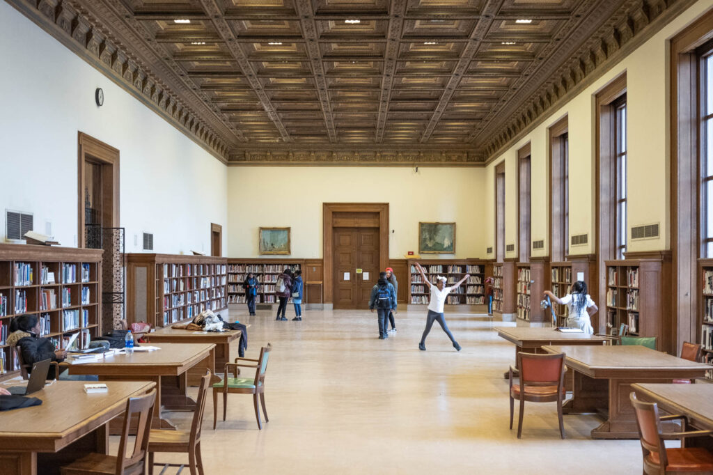 Interior of a Beaux-Arts library reading room with large rectangular windows and a wood-panelled ceiling. Children are playing in the space.