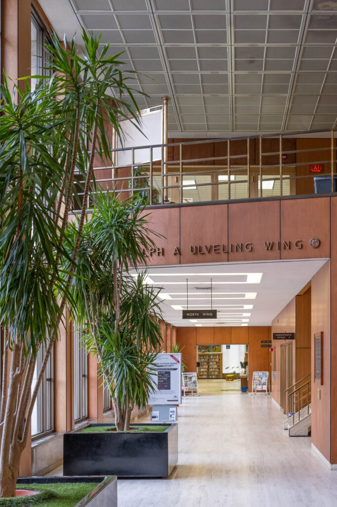 Interior of a Modernist library with wooden wall panels and large potted plants. The image looks towards a hallway marked "North Wing" and "Ralph A. Ulveling Wing".