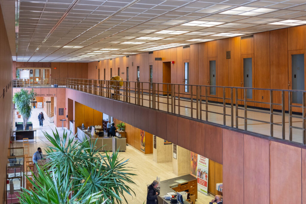 Interior of a Modernist library with wooden wall panels, showing a mezzanine opening to a long rectangular atrium where patrons are milling. Large potted plants stand in the atrium.
