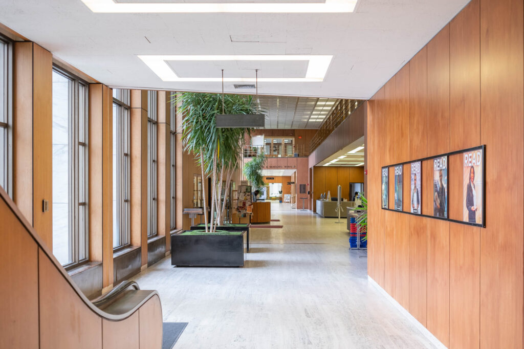 Interior of a Modernist library with wooden wall panels, large indoor plants and an escalator to the left.