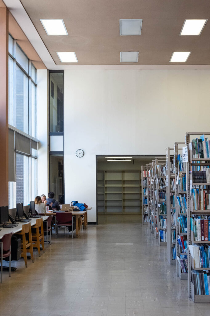 Interior of a modernist library, in beige and ochre tones. There are bookshelves to the right and students working at tables to the left.