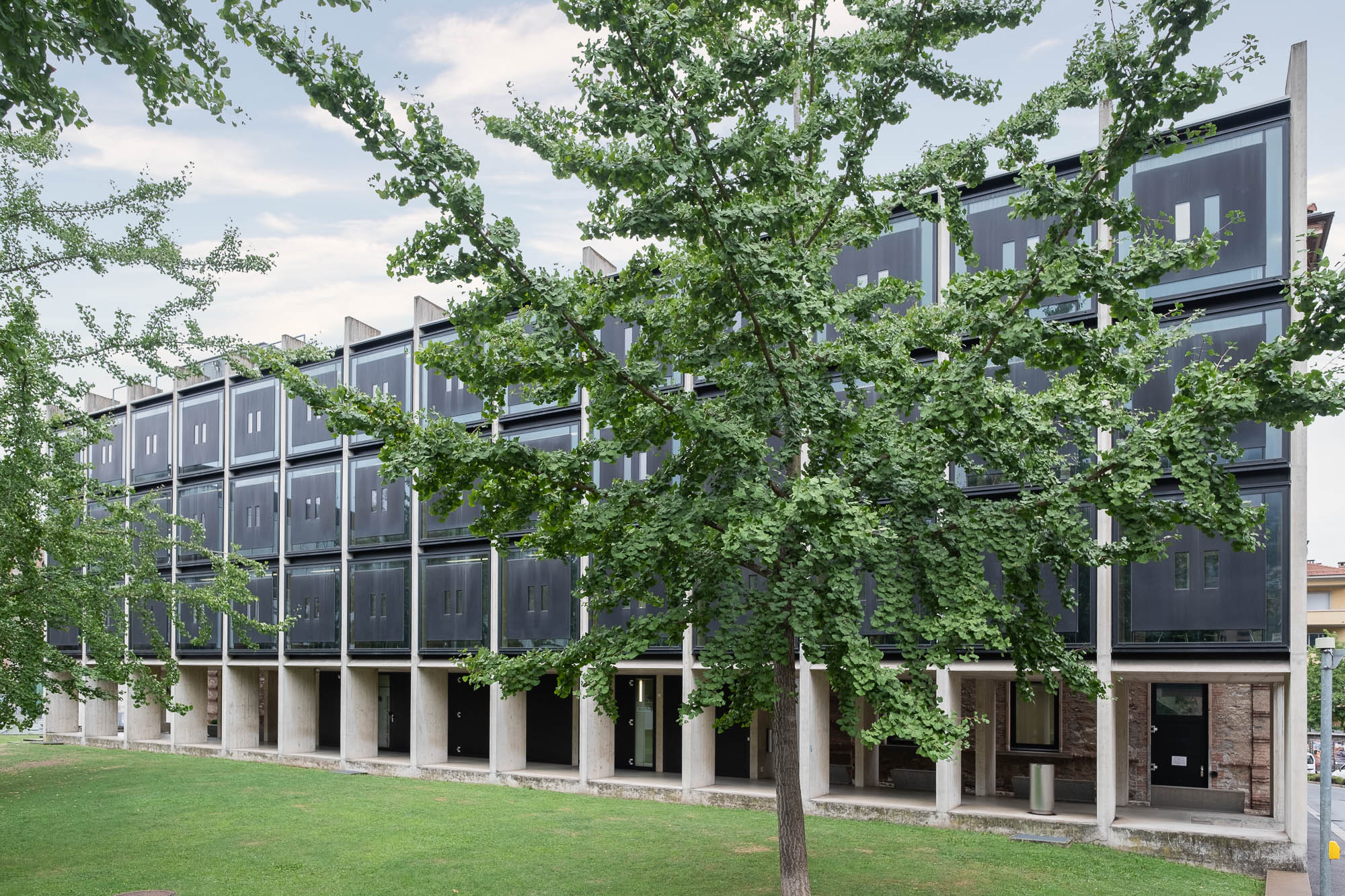 Exterior of the USI library in Lugano, Switzerland, showing its contemporary east facade, which resembles a lattice of concrete elements supporting dark metal boxes. There are trees and a lawn at the front. To the right, the street can be seen fronting the building.