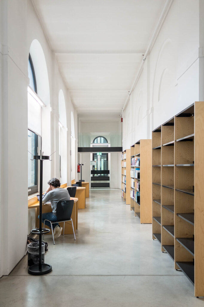 Interior of a refurbished neobaroque building, showing a corridor with vaulted windows to the left, fronted by small work tables. A person sits at one of the tables. There are wooden bookcases to the right.