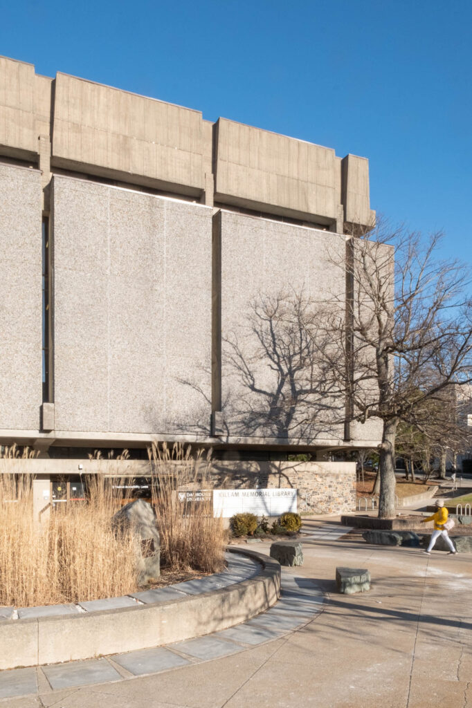 Exterior of a Brutalist library with a facade of large textured concrete panels between which are narrow vertical slit windows. A person is walking towards the library from the edge of the image.