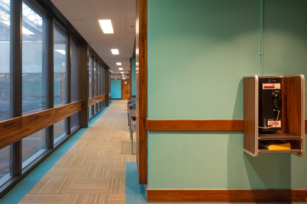 Interior view of a library, showing a corridor alongside an glass facade opening to a covered atrium. To the right is an older-looking phone booth.