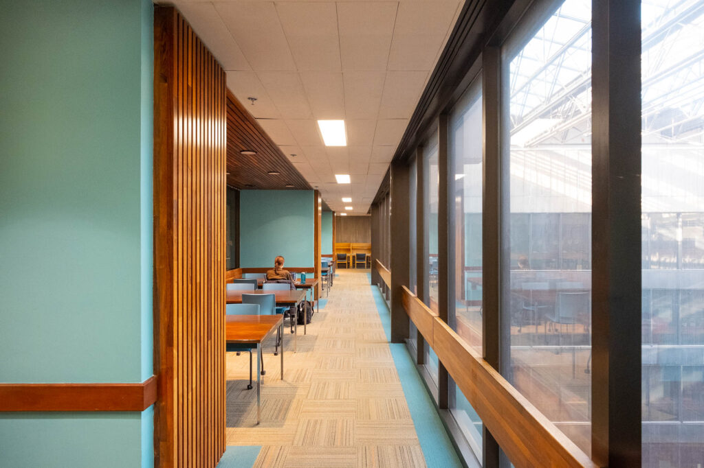 Interior view of a library, showing a corridor alongside an glass facade opening to a covered atrium. To the left are rows of wooden tables. A person is seen from the back sitting at one of the tables.