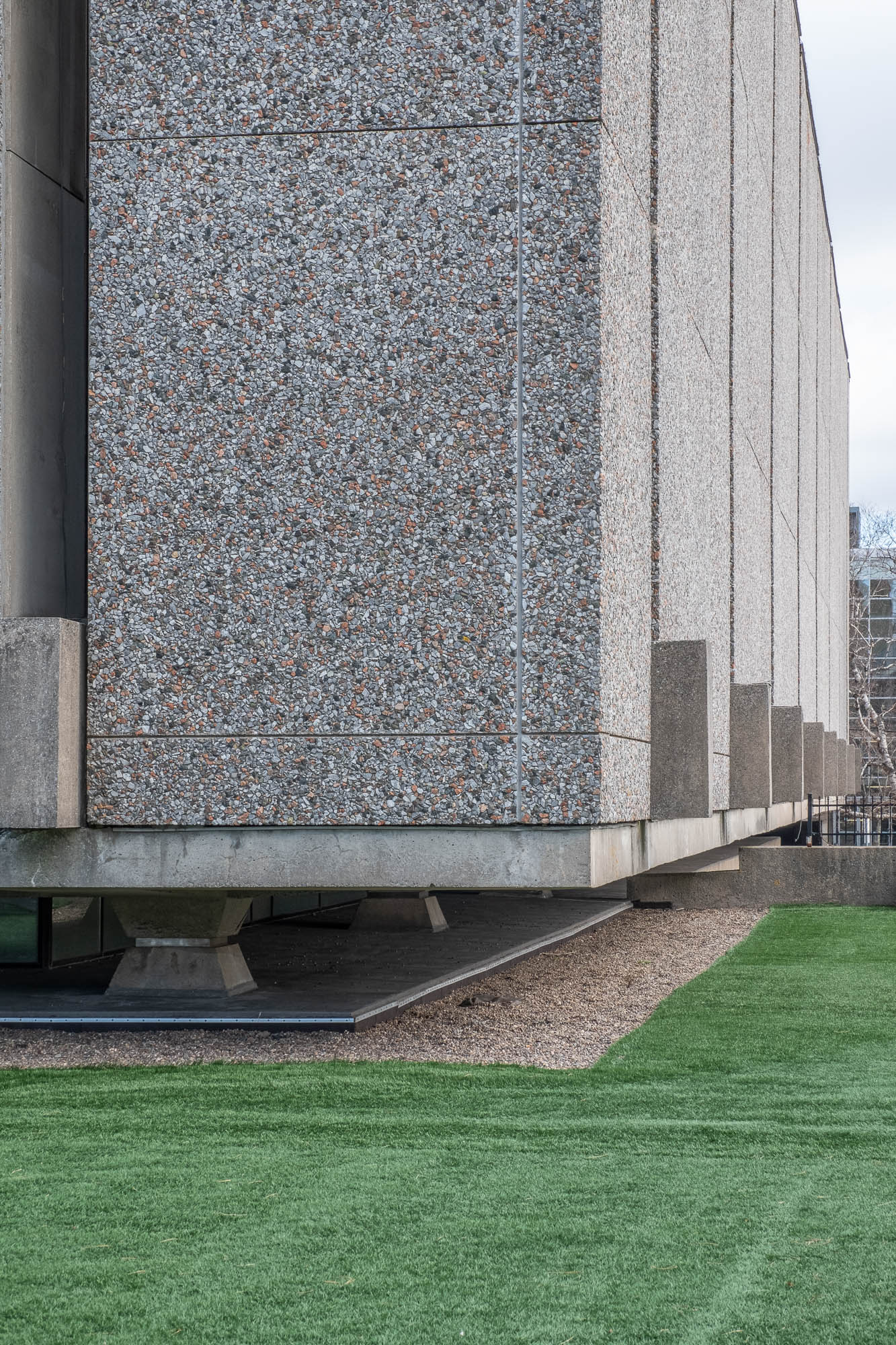 Exterior of a Brutalist building, close-up view of a corner of the building. We see details of textured concrete panels. The building is raised a few feet above ground, which is covered in artificial grass.