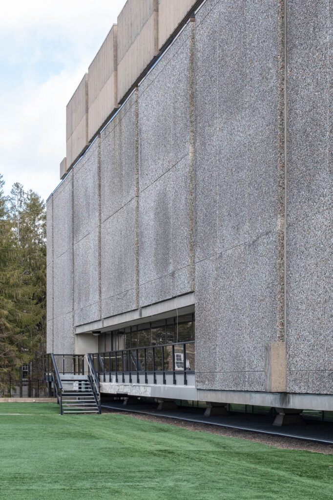 Exterior of a Brutalist library with a facade of large textured concrete panels between which are narrow vertical slit windows.