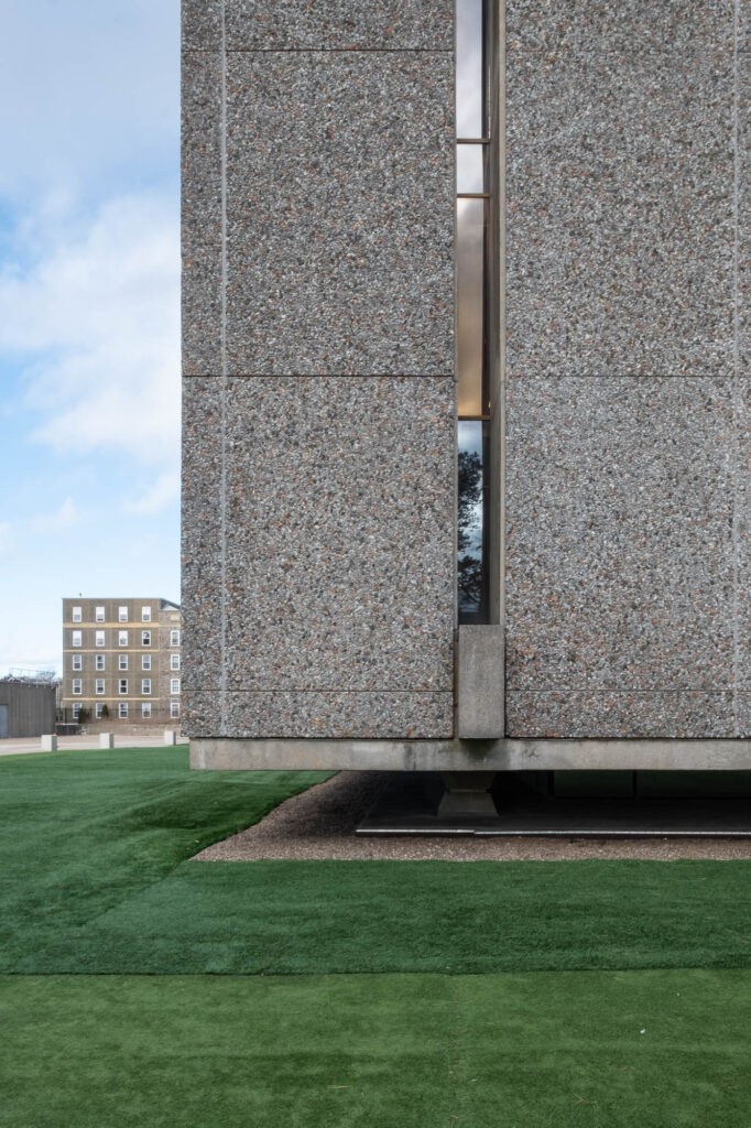 Exterior of a Brutalist building, close-up view of a corner of the building. We see details of textured concrete panels. The building is raised a few feet above ground, which is covered in artificial grass.