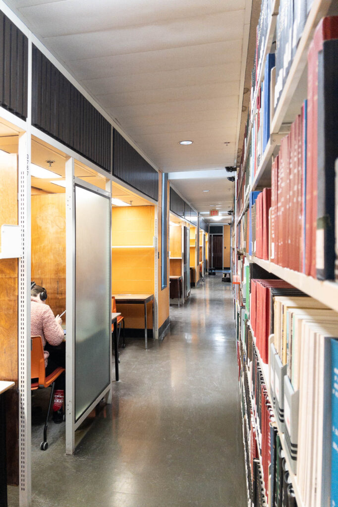 View of a corridor inside a library, with a high bookshelf on the right filled with large volumes and work cubicles to the left. One of the cubicles is partly covered by a frosted glass window. A person wearing headphones can be seen through the gap working at the cubicle.