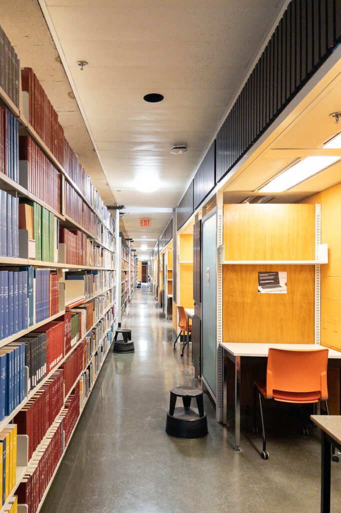 View of a corridor inside a library, with a high bookshelf on the left filled with large volumes and work cubicles to the right. Stepping stools are spread around the corridor.