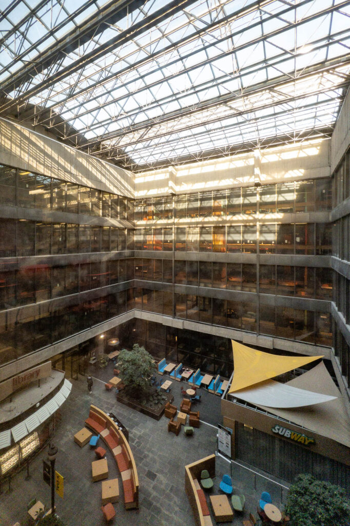 View of a covered atrium with glassed facades all around it. The bottom of the atrium is taken by two snack bars, chairs and booths, as well as large interior trees.