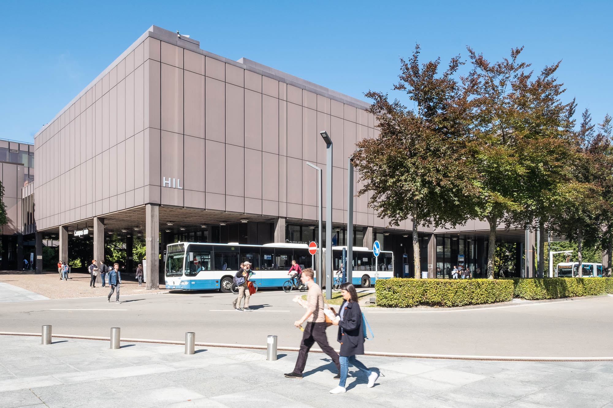 Exterior of a modern library. It is a rectangular box on stilts, with a blind facade of reddish metal panels. A blue and white city bus is parked in front of the building and people are milling on the street in front. The building bears the letters HIL.