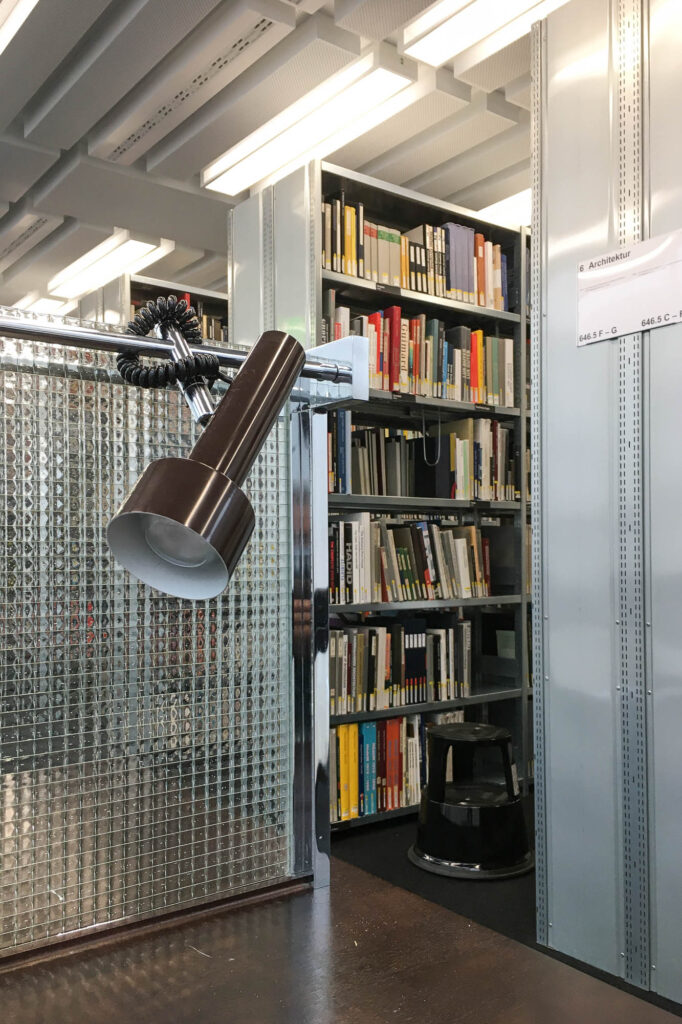 Interior of a library, showing metal bookshelves in the background and a table with a light in the foreground.
