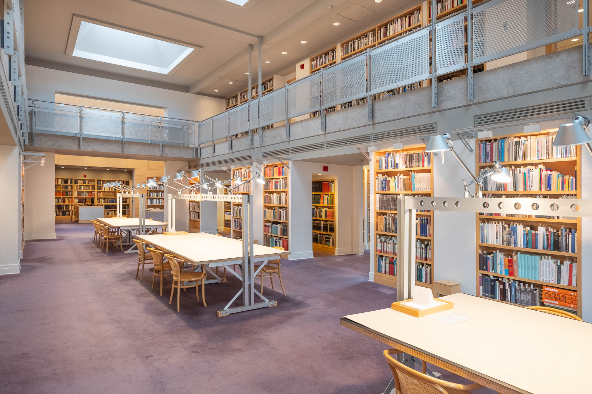 Interior of a contemporary library. It is a symmetrical room surrounded by a mezzanine and with large work tables in the middle.