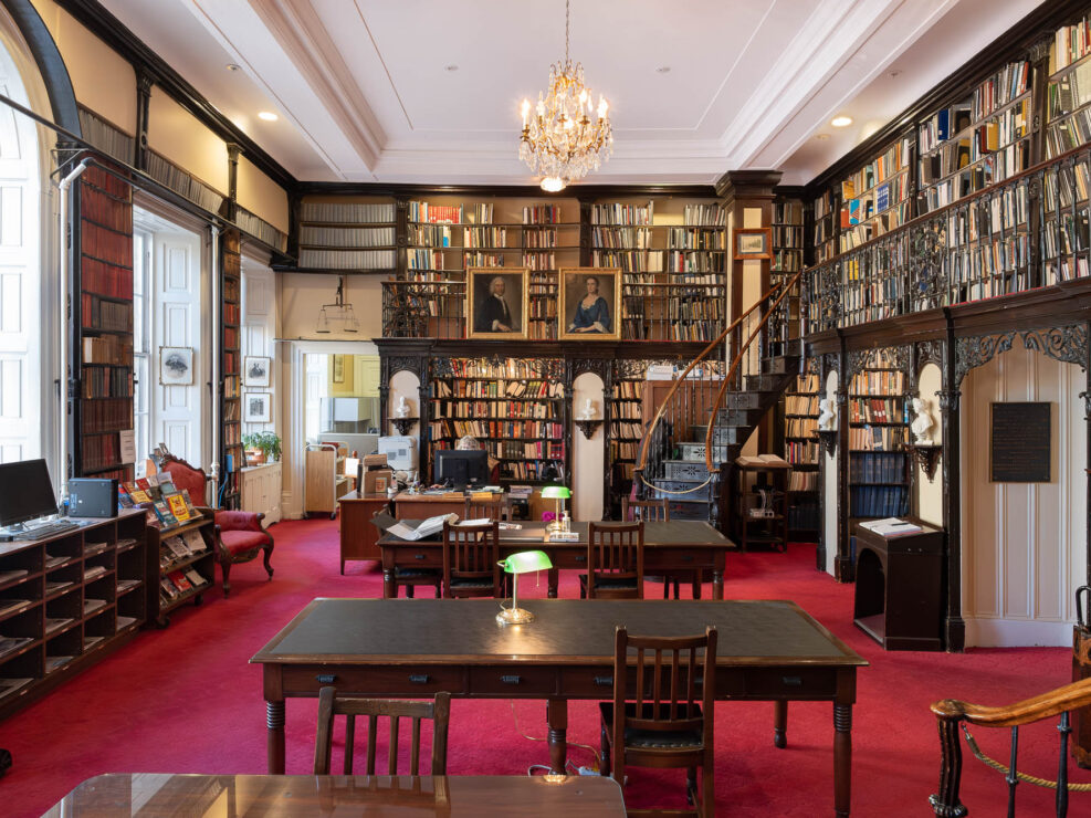 Interior of a classical library with dark wood shelves adorned with intricate black iron railings. plaster busts on consoles and oil portraits. A chandelier hangs from the ceiling.