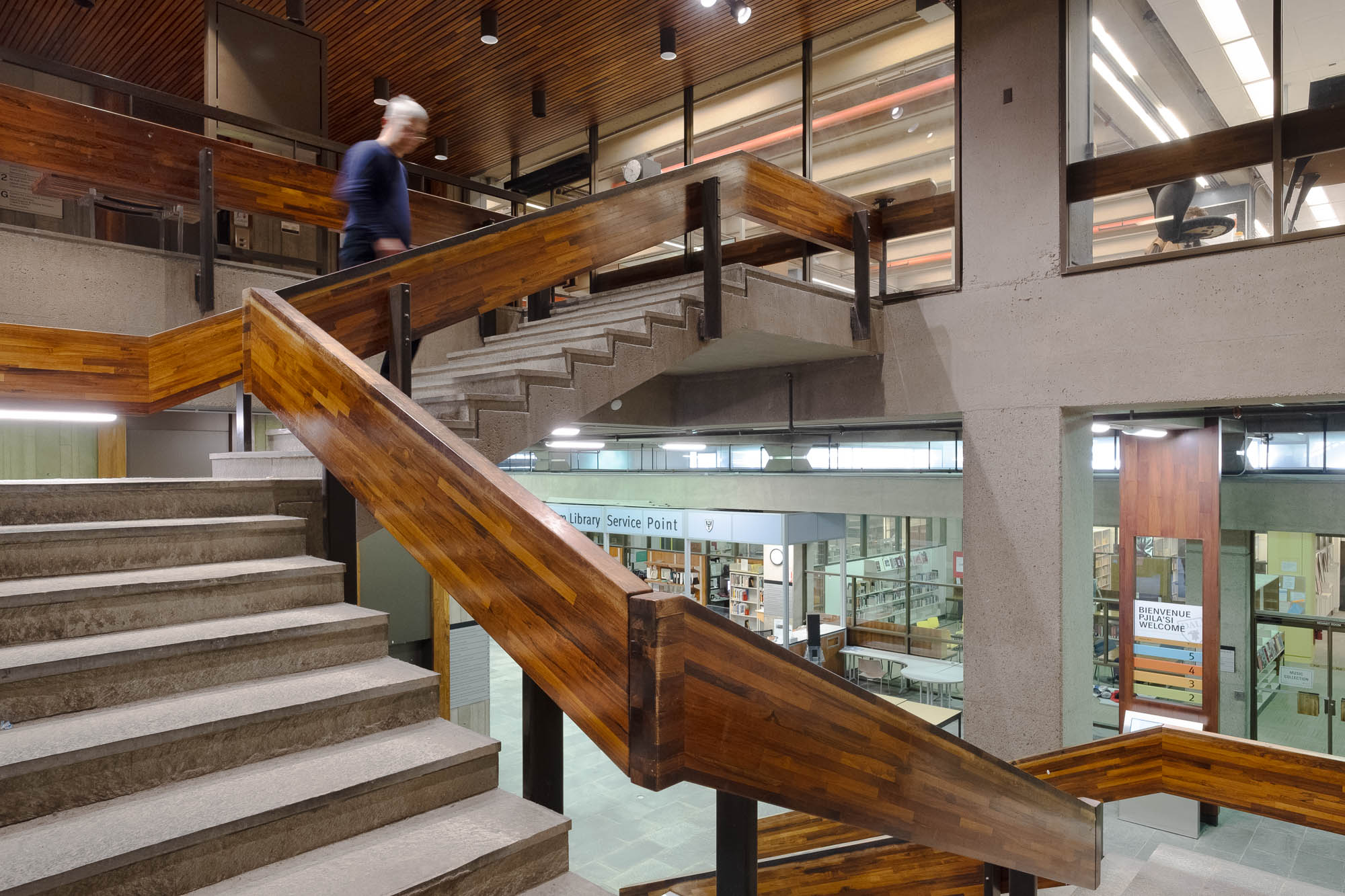Interior of a Brutalist library, showing a staircase. A figure is climbing the stairs, blurred by movement. On the main ground below is a library welcome desk with the words "Library service point" on it. To the right is a sign reading "Bienvenue, Pjila'si, Welcome".