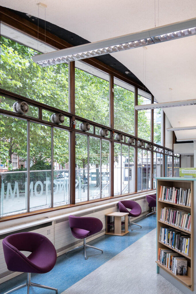 Interior of a modern library, with a gently curving ceiling and a large window. Colourful contemporary furniture line the window.