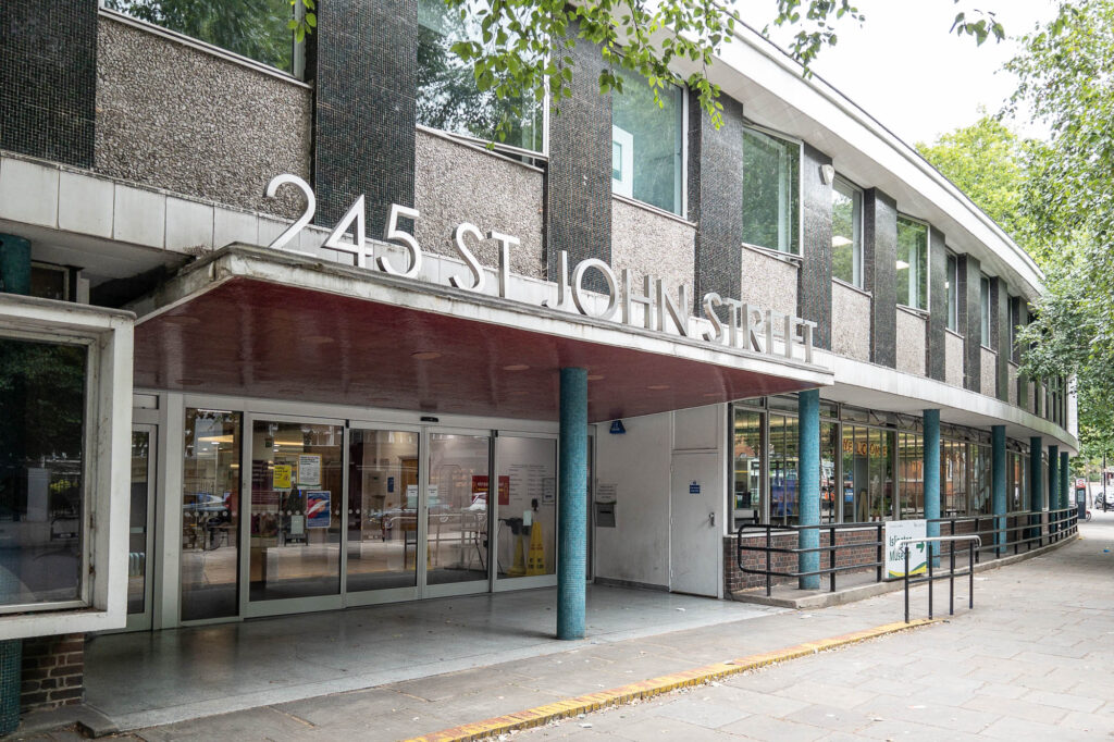 Exterior of a Modernist library, a gently curving facade clad in teal, burgundy and black ceramic tiles with concrete aggregate panels. At the forefront is a porte cochère with steel letters reading "St John Street".
