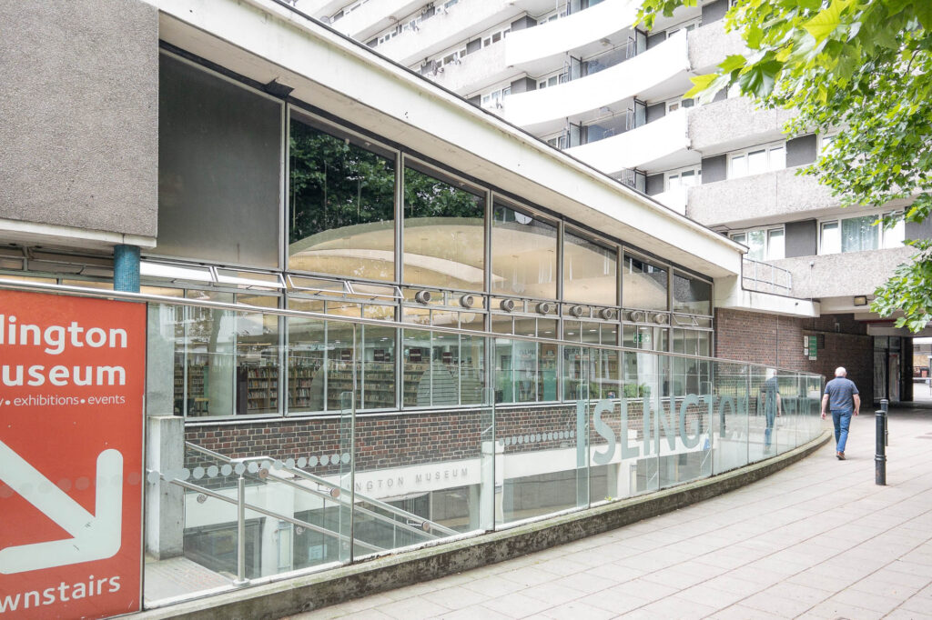 Exterior of a Modernist library, with a gently sloping flat roof, and a glass, brick and concrete facade. Through the glass, the curve of the interior ceiling is visible.