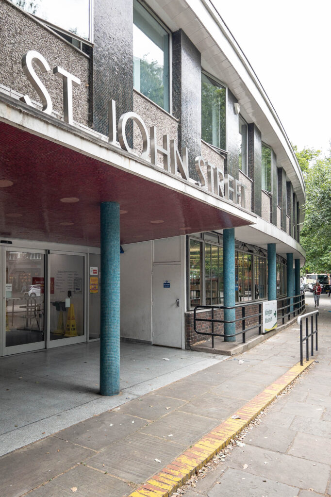 Exterior of a Modernist library, a gently curving facade clad in teal, burgundy and black ceramic tiles with concrete aggregate panels. At the forefront is a porte cochère with steel letters reading "St John Street".