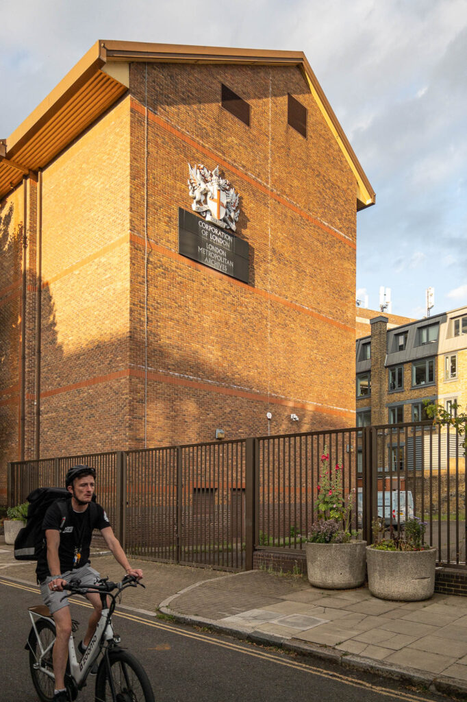 Exterior of a tall brick building with an asymmetric pitched roof. There are no windows on the facade, but two small ventilation grilles. A crest and a sign with the words "Corporation of London. London Metropolitan Archives" is affixed to the wall. A man riding a bicycle is seen in the forefront.