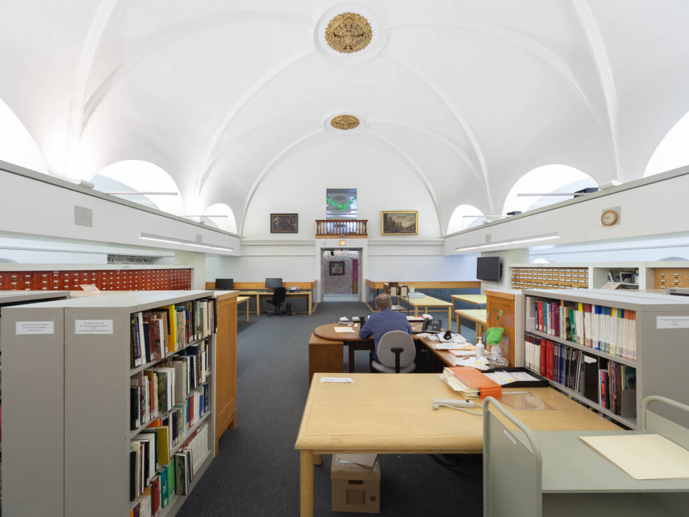 Interior of a library in a converted chapel, wide-angle image of a reading room and librarian's workspace, with low metal shelves and cabinets, tables, and card catalogues. The vaulted ceiling is white with golden details.