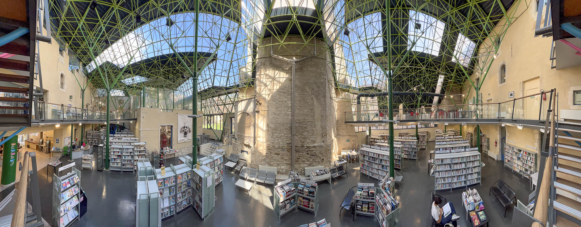 Interior of a library inside a converted former hospital, a high glass roof on metal stilts covers an area bound by original stone walls. The metal stilts branch out as they rise and are painted green, recalling tree trunks.