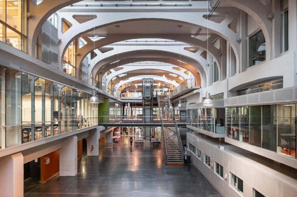 A spacious former industrial building interior with high arched ceilings, glass walls, and open walkways. A central staircase leads to upper levels, with workspaces and offices visible on both sides. The area is well-lit and empty.