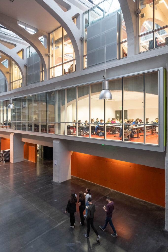 A group of people walk inside a spacious former industrial building with high ceilings and large windows. Above them, a classroom filled with students is visible through glass walls. The interior features white arches and orange accent walls.