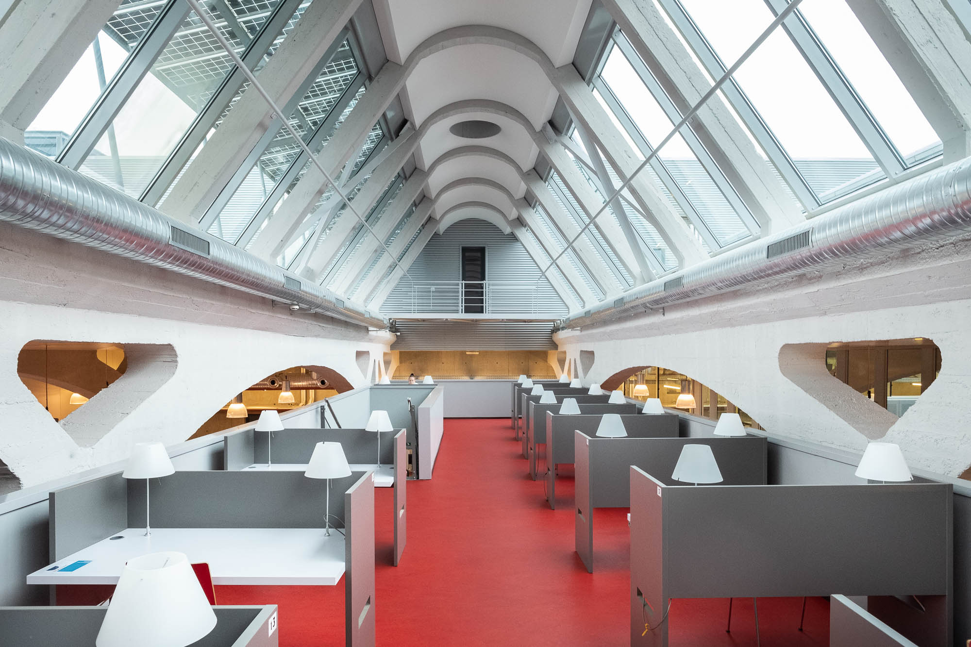 A modern workspace with high vaulted ceilings and large skylights. Rows of gray cubicles with white desk lamps are arranged on a bright red floor, with a mezzanine visible at the far end.