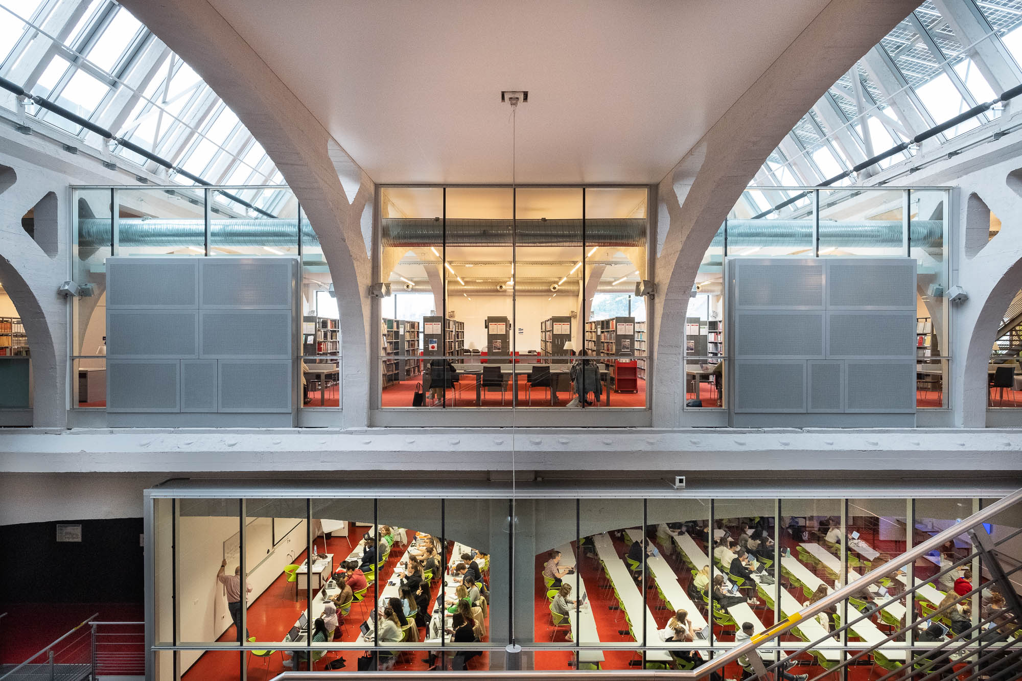 Interior view of a university campus building in a former industrial space. The upper level has bookshelves and people studying, while the lower level holds classrooms with students seated at long tables, seen through glass walls. The architecture features large white arches.