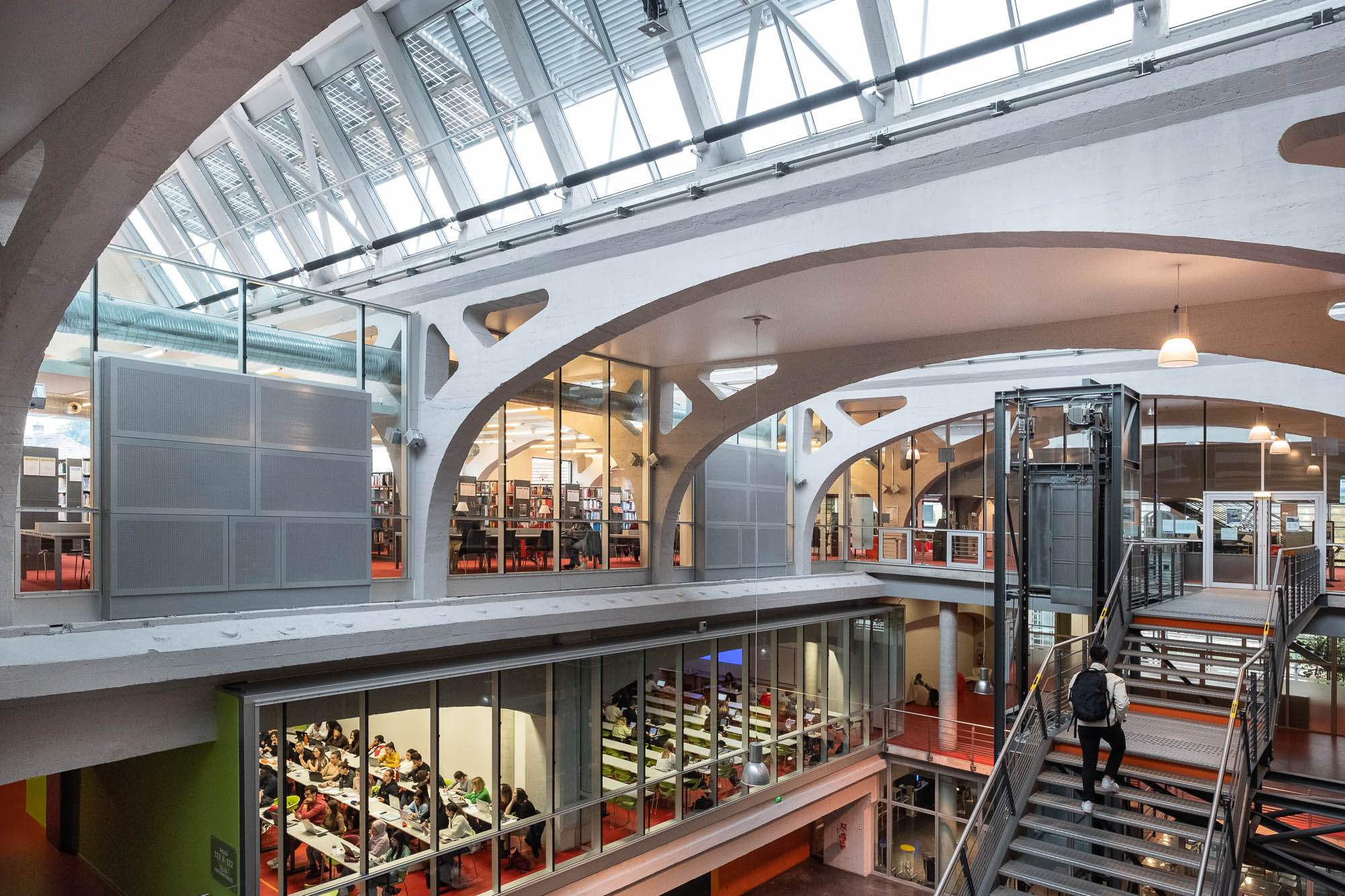Interior view of a university campus building in a former industrial space. The upper level has bookshelves and people studying, while the lower level holds classrooms with students seated at long tables, seen through glass walls. To the right are metal stairs on which a person is climbing. Large concrete arches support a shed roof overhead.