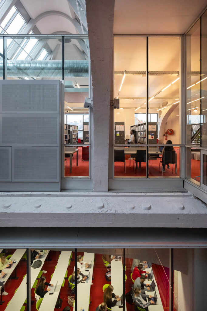 Interior view of a university campus building in a former industrial space. The upper level has bookshelves and people studying, while the lower level holds classrooms with students seated at long tables, seen through glass walls.