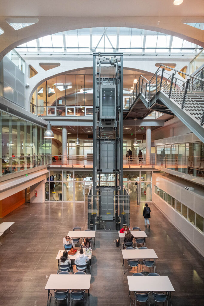 Interior view of a university campus building in a former industrial space. The upper level has bookshelves and people studying, while the lower level holds classrooms with students seated at long tables, seen through glass walls. The open space below has tables with people seated at them. At the back is an elevator shaft and to the right are metal stairs. Large concrete arches support a shed roof overhead.