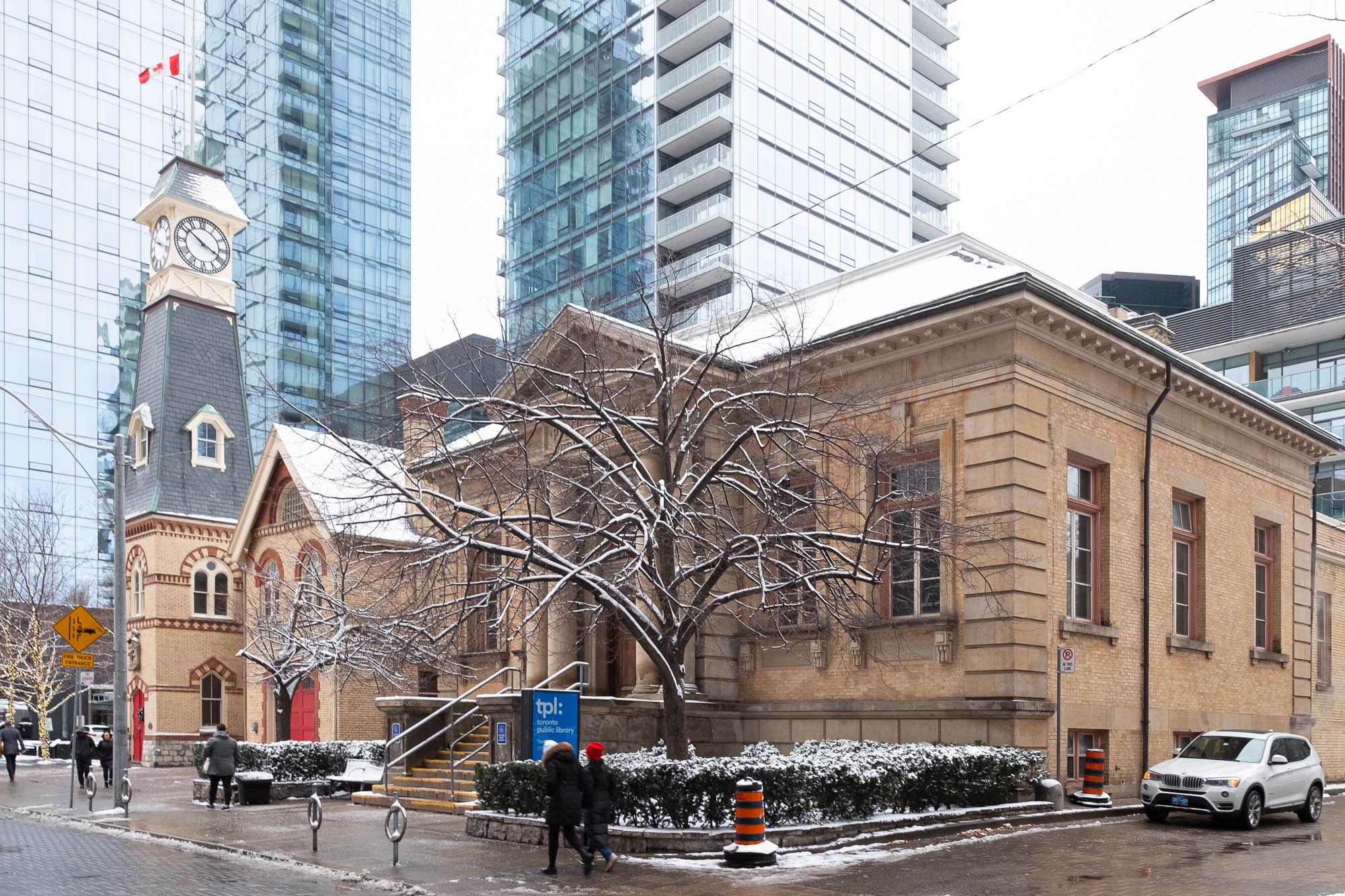 Exterior of a small Neoclassical library. It is a symmetrical, single-story brick building. The entrance door is framed by a pediment supported by four columns on top of a flight of stairs. In front of the library, a modern sign reads "tpl: Toronto Public Library". There is snow on the ground and the sky is overcast. Behind the library is a brick fire station flanked by a clock tower flying a Canadian flag. The ensemble is surrounded by tall glass towers.