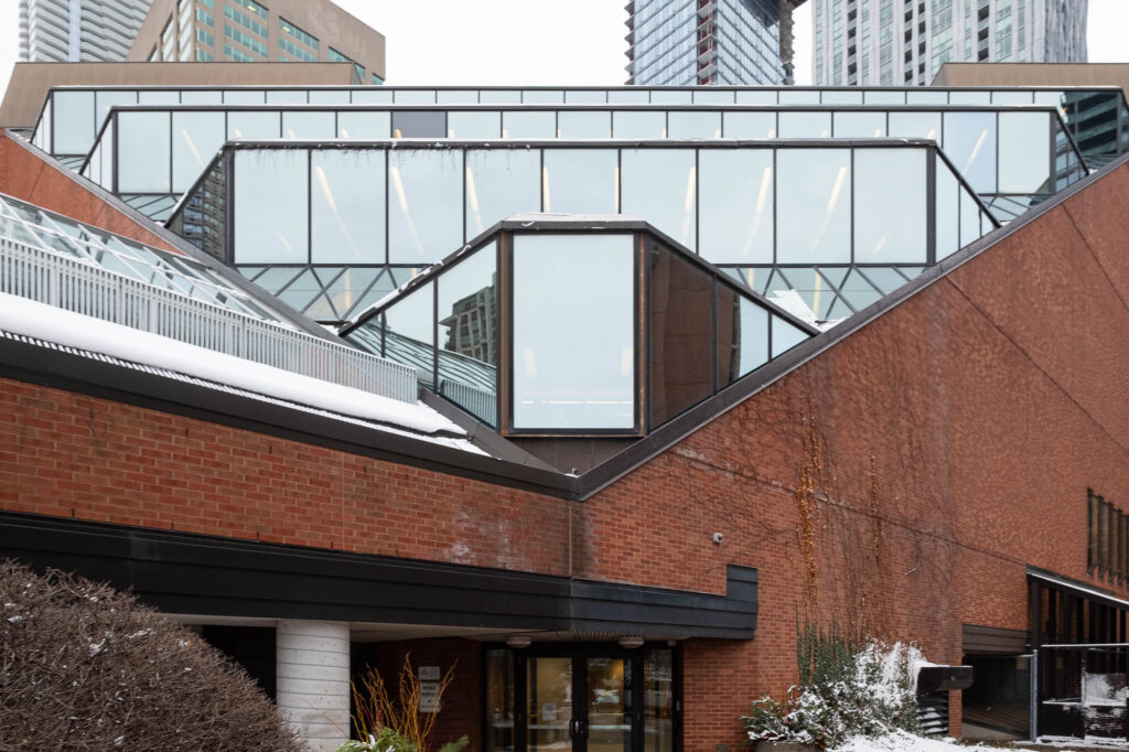 Geometric architecture with a mix of brick and glass elements. A prominent triangular glass structure extends from the building facade. Snow is visible on some surfaces, with city buildings in the background.