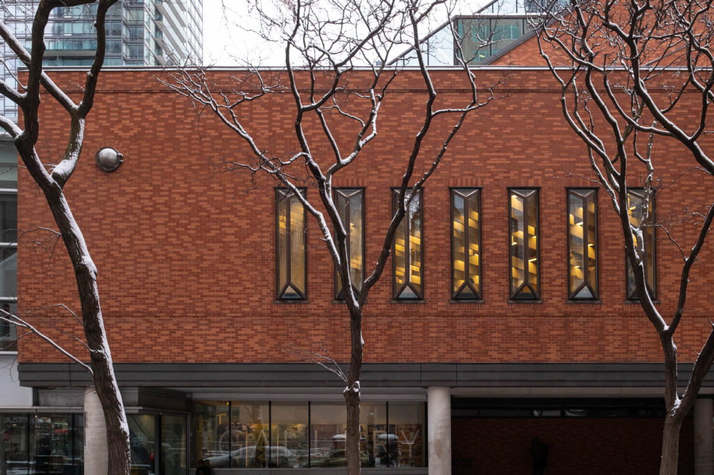 A modern building with a red brick facade featuring five tall, narrow windows. Leafless trees with snow-dusted branches frame the view. The lower level has glass walls and a visible entrance. Urban high-rise buildings are seen in the background.