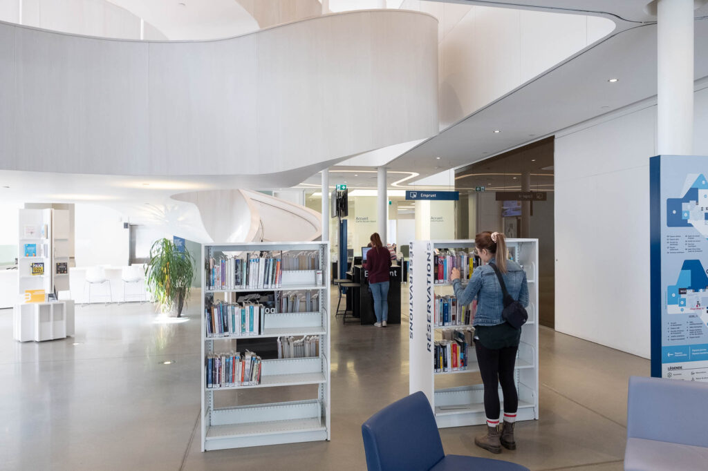 Interior of a contemporary library entrance area, with small holds bookshelves and checkout machines. A spiral staircase is seen in the background.