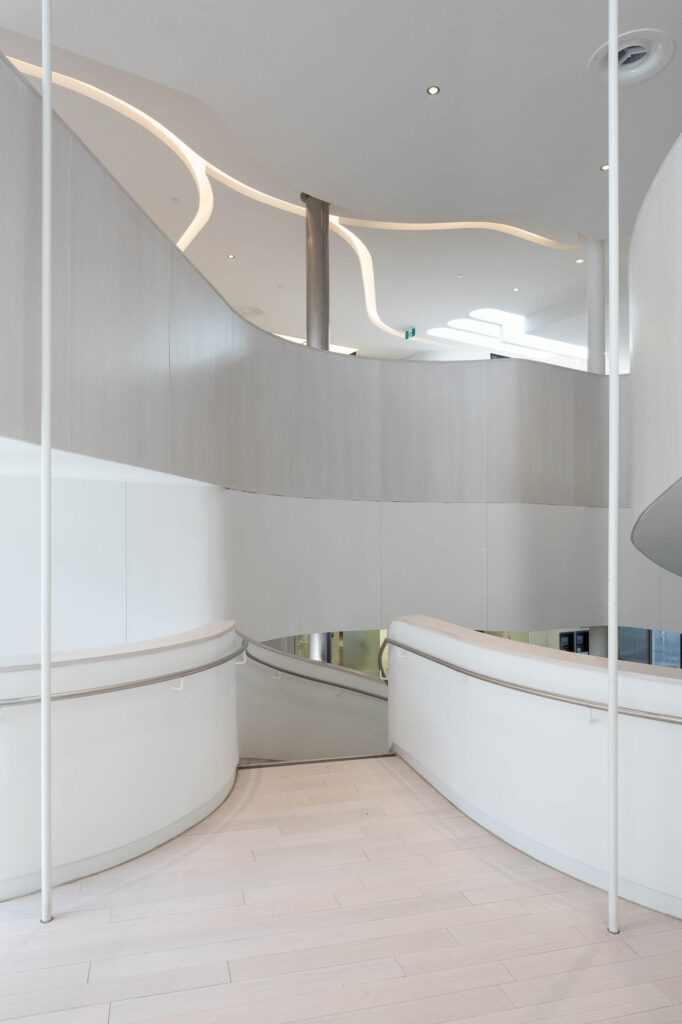 Interior of a contemporary library, showing a curving staircase in white and cream colours. Light filters down from curving slits in the ceiling.